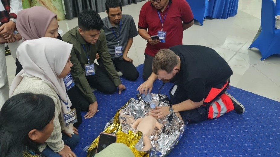 Training participants encircling an instructor who show how to do CPR for infants by practicing the method on a baby doll.